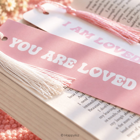 Two pink and white book bookmarks with tassels displayed on open book pages. One bookmark is pink with white text reading 'YOU ARE LOVED' and the other is white with pink text reading 'I AM LOVED'.
