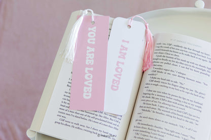 Two pink and white book bookmarks with tassels displayed on open book pages. One bookmark is pink with white text reading 'YOU ARE LOVED' and the other is white with pink text reading 'I AM LOVED'.