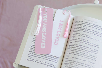 Two decorative bookmarks shaped like book pages, one pink and one white, both with tassels and the words 'YOU ARE LOVED' and 'I AM LOVED' printed vertically, resting on an open book.