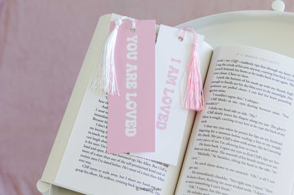 A pair of white book bookmarks with pink tassels and pink lettering, shown bookmarking an open book on a soft pink background. One bookmark reads 'YOU ARE LOVED' and the other reads 'I AM LOVED'.