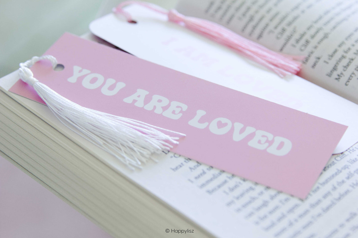 A pink paper bookmark with white tassels and the words 'YOU ARE LOVED' in white lettering, resting on an open book alongside other bookmarks.
