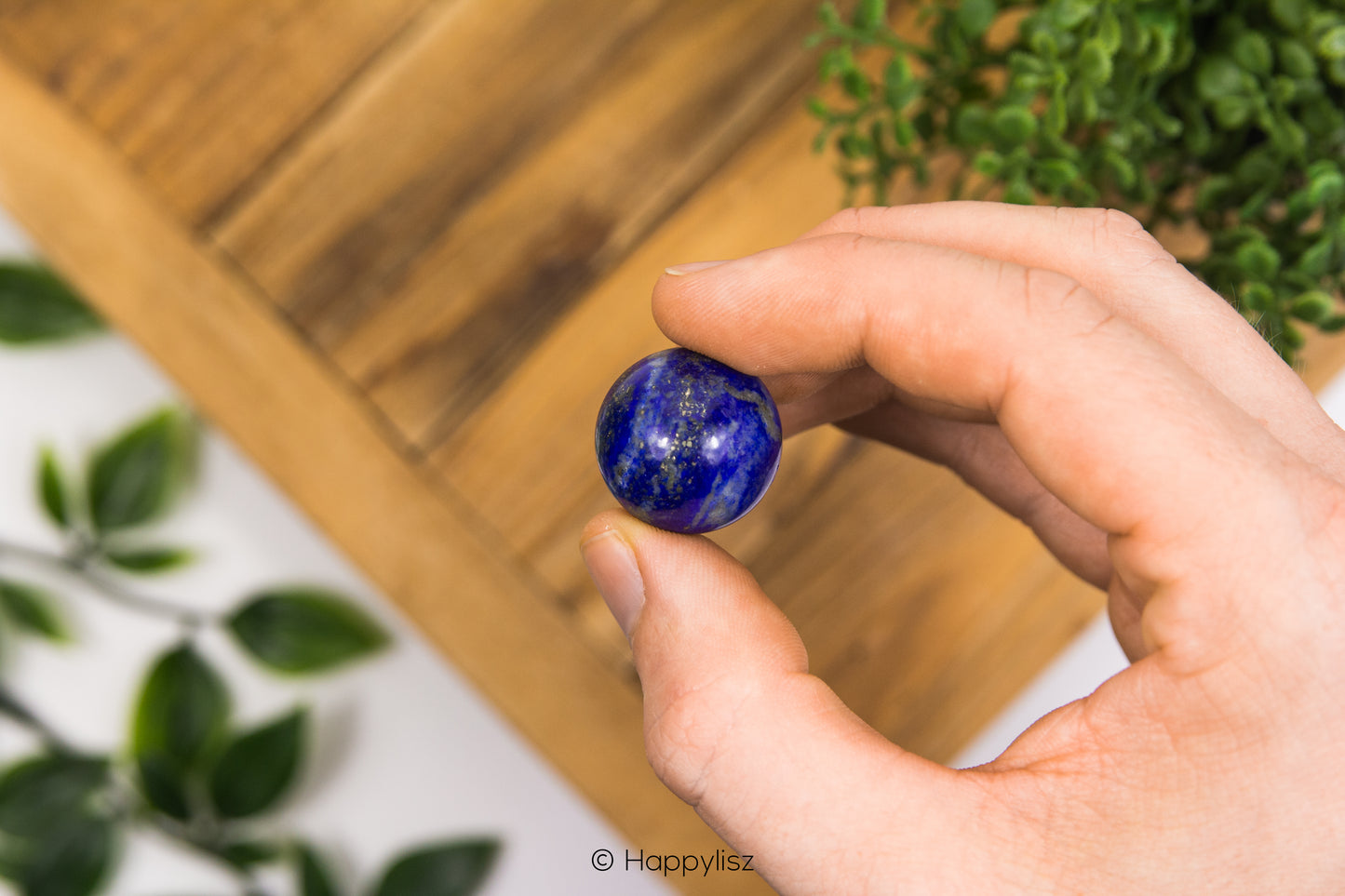 Hand holding a blue gemstone with a blurred background of green plants and wooden surface