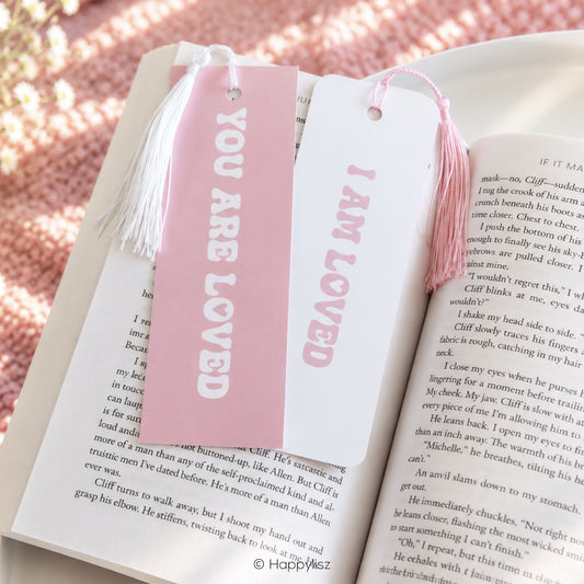 Two pink and white book bookmarks with tassels displayed on open book pages. One bookmark is pink with white text reading 'YOU ARE LOVED' and the other is white with pink text reading 'I AM LOVED'.